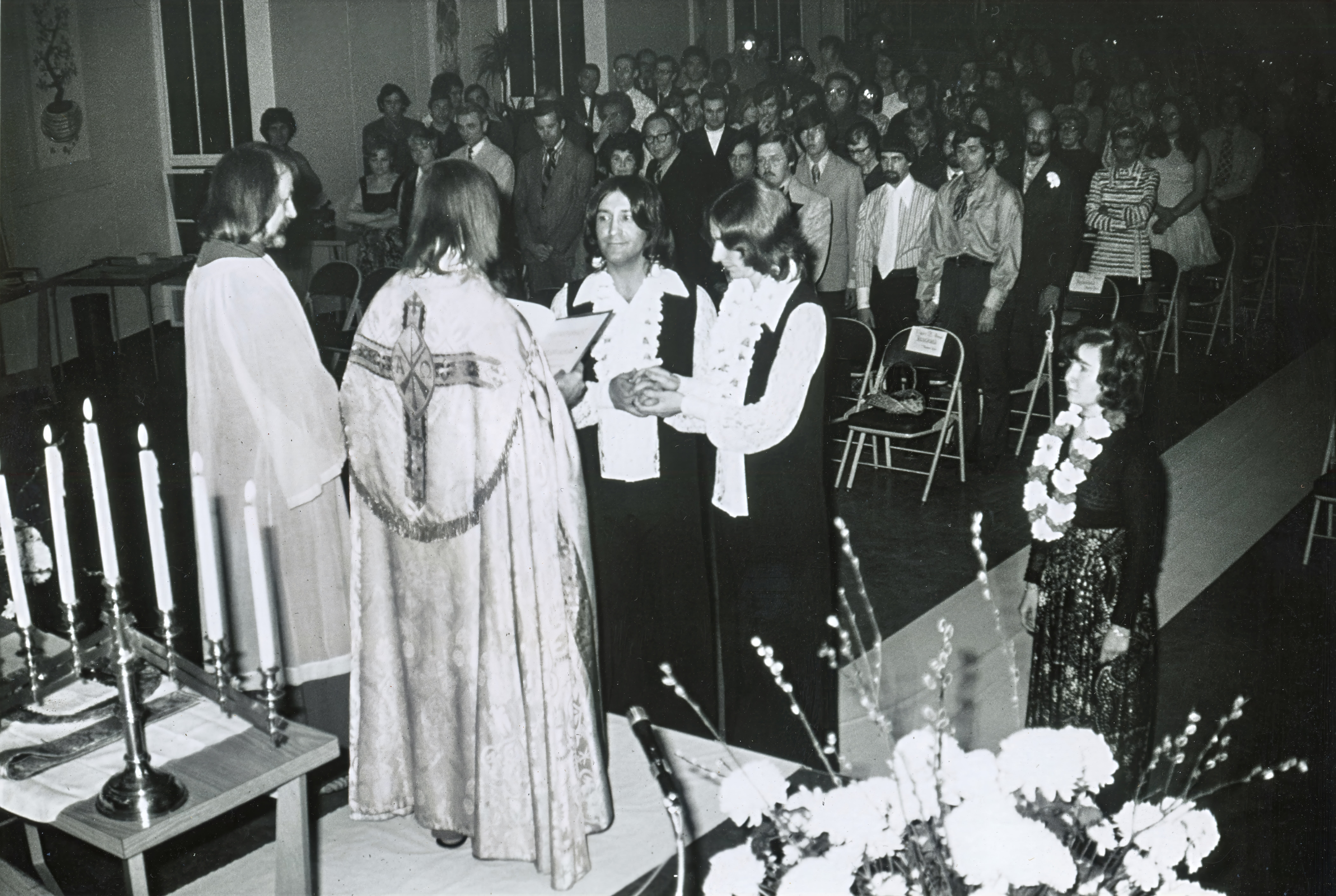 This photograph captures one of the Church of the Beloved Disciple's Holy Union ceremonies, officiated by Fr. Robert Mary Clement before a large congregation at an unidentified location. Following a liturgy modeled closely on the Christian marriage rite, the couple exchanged vows and rings as the community gathered to witness and bless their covenant. Throughout the 1970s, these public ceremonies offered LGBTQ couples a sacramental affirmation of their relationships decades before civil marriage equality became a reality in the United States.