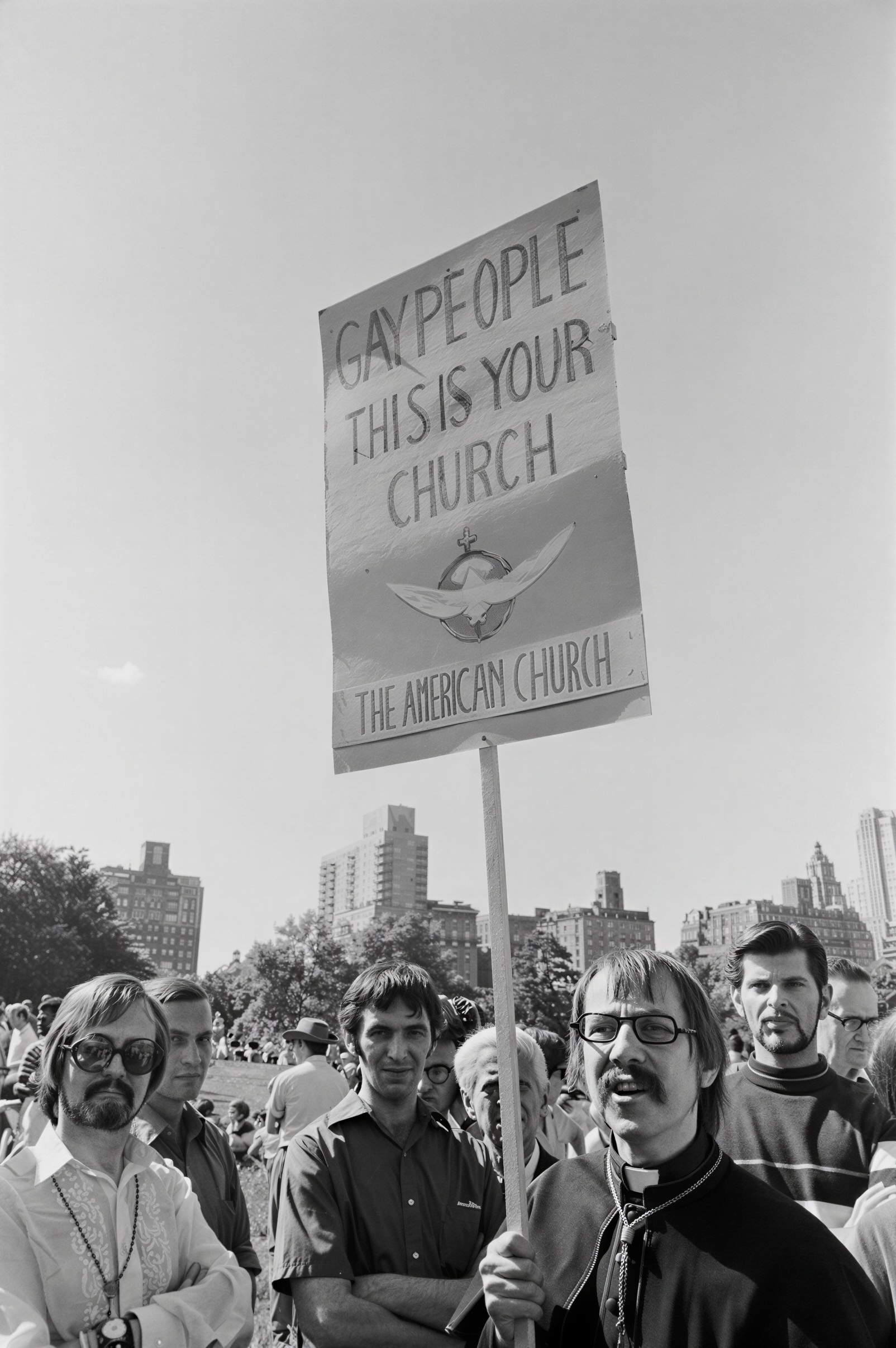 Robert M. Clement and John Darcy Noble stand in Sheep Meadow following the first Christopher Street Liberation Day March on June 28, 1970, beneath a hand-painted sign proclaiming, "Gay People, This Is Your Church." Earlier that day, Clement carried this sign in the march while Noble distributed leaflets announcing their forthcoming inaugural public Mass. Carried at the first anniversary march commemorating the Stonewall uprising, the sign publicly proclaimed the Church of the Beloved Disciple’s affirming sacramental ministry for lesbian and gay people ahead of its first public Mass on July 19, 1970. This restored photograph preserves one of the earliest surviving public images of Clement and Noble's ministry and documents a pioneering declaration that LGBTQ Christians could participate fully in the sacramental life of the Church without concealing their identities.