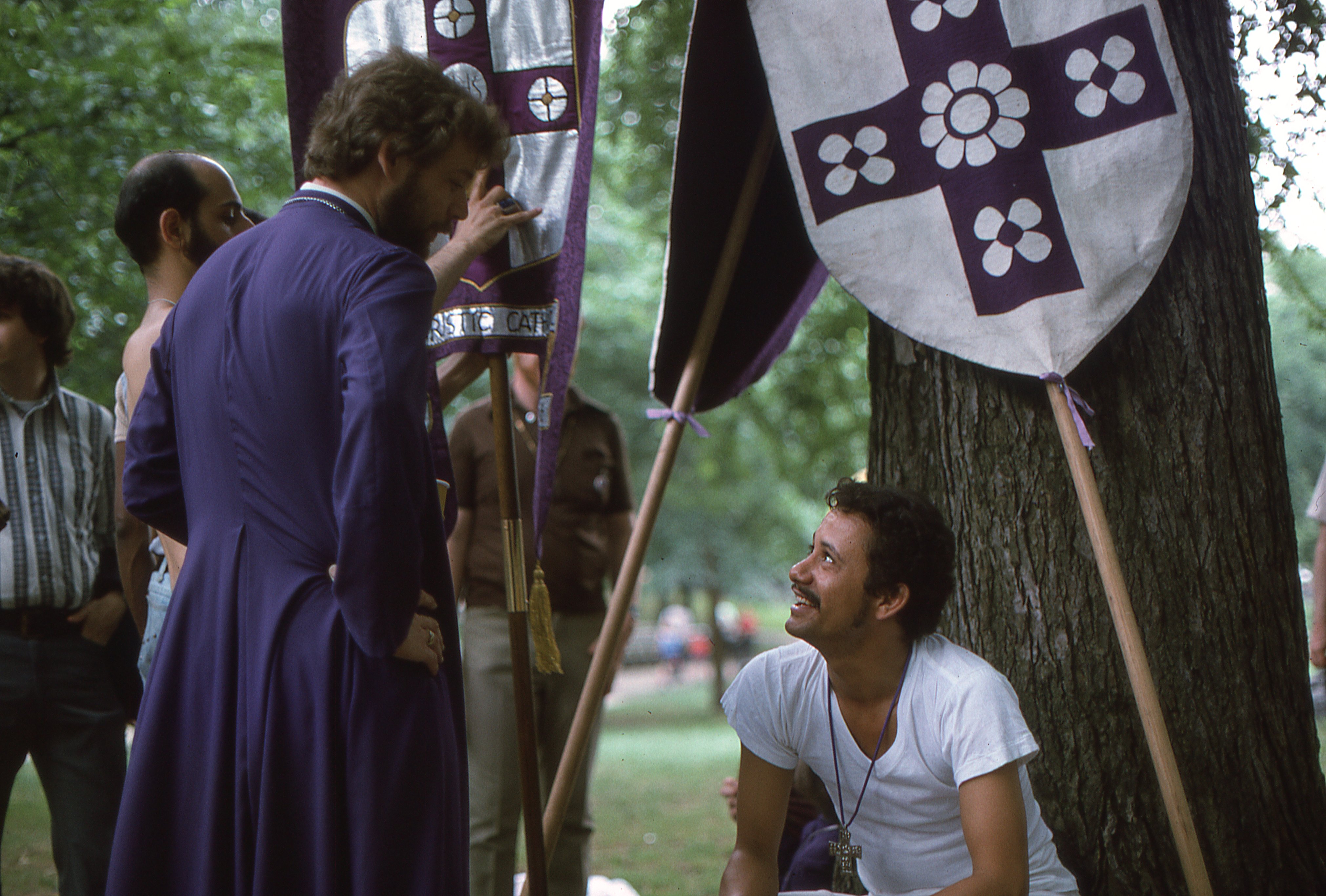Fr. Leo speaks with a member of the Church of the Beloved Disciple in Sheep Meadow following the Fifth Annual Christopher Street Liberation Day March on June 30, 1974. Surrounding them are the church's processional banners and heraldic standard, while the congregant wears a distinctive silver cross designed by Fr. Leo and adopted by members of the congregation as a symbol of their shared faith and community.