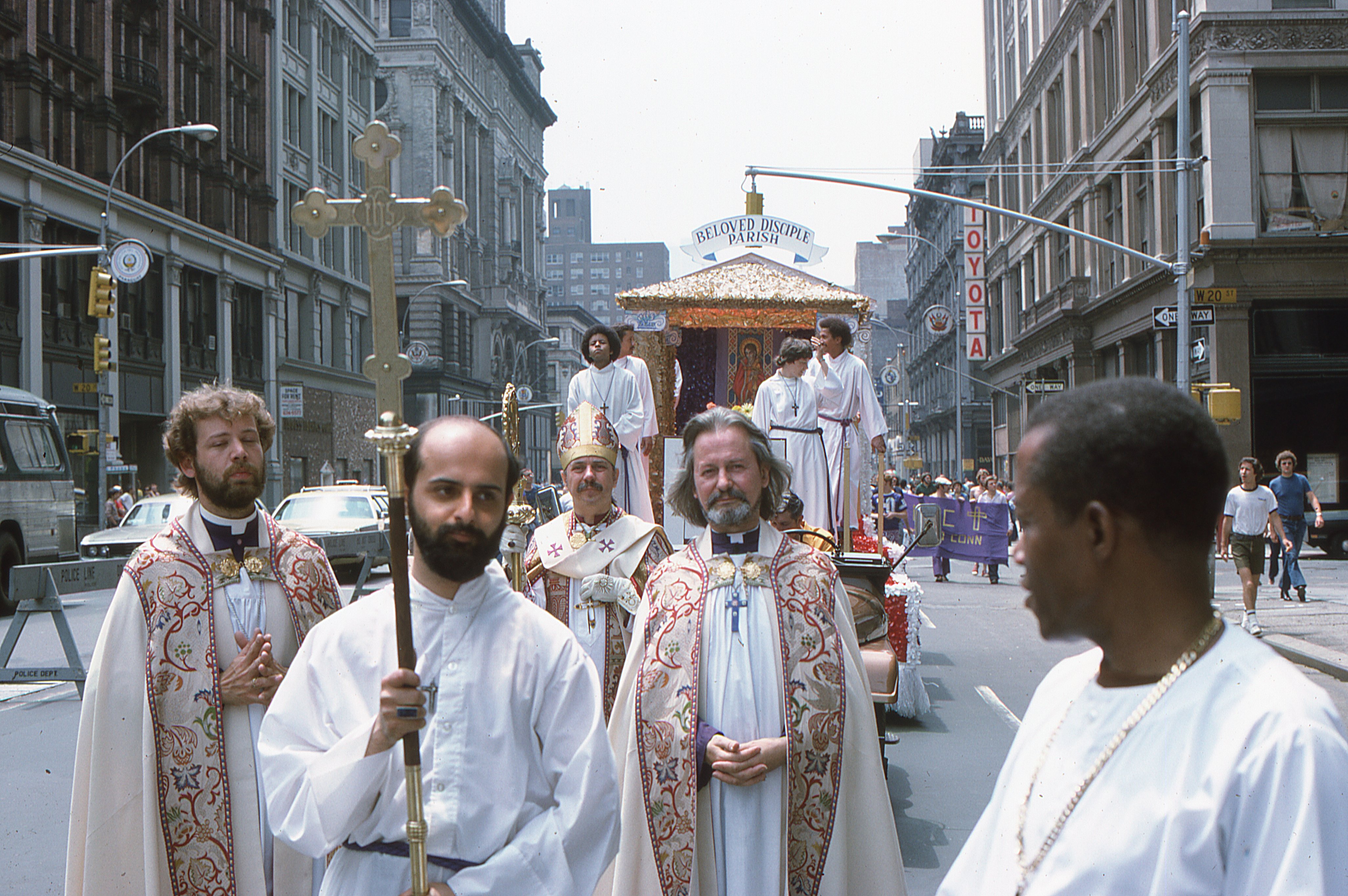 Clergy of the Church of the Beloved Disciple process ahead of the congregation's float during the Sixth Annual Gay Pride Day March (Gay Liberation Day) in New York City on June 29, 1975. From left to right are Fr. Leo (partially visible), Fr. John (center), and Bishop Robert Mary Clement (wearing the mitre), accompanied by unidentified acolytes and cross-bearers. Behind them, the Church of the Beloved Disciple's float, designed as a processional chapel, affirmed the congregation's conviction that LGBTQ Christians belonged fully within the sacramental life of the Church and brought liturgical worship into one of the nation's most visible public celebrations of gay liberation.