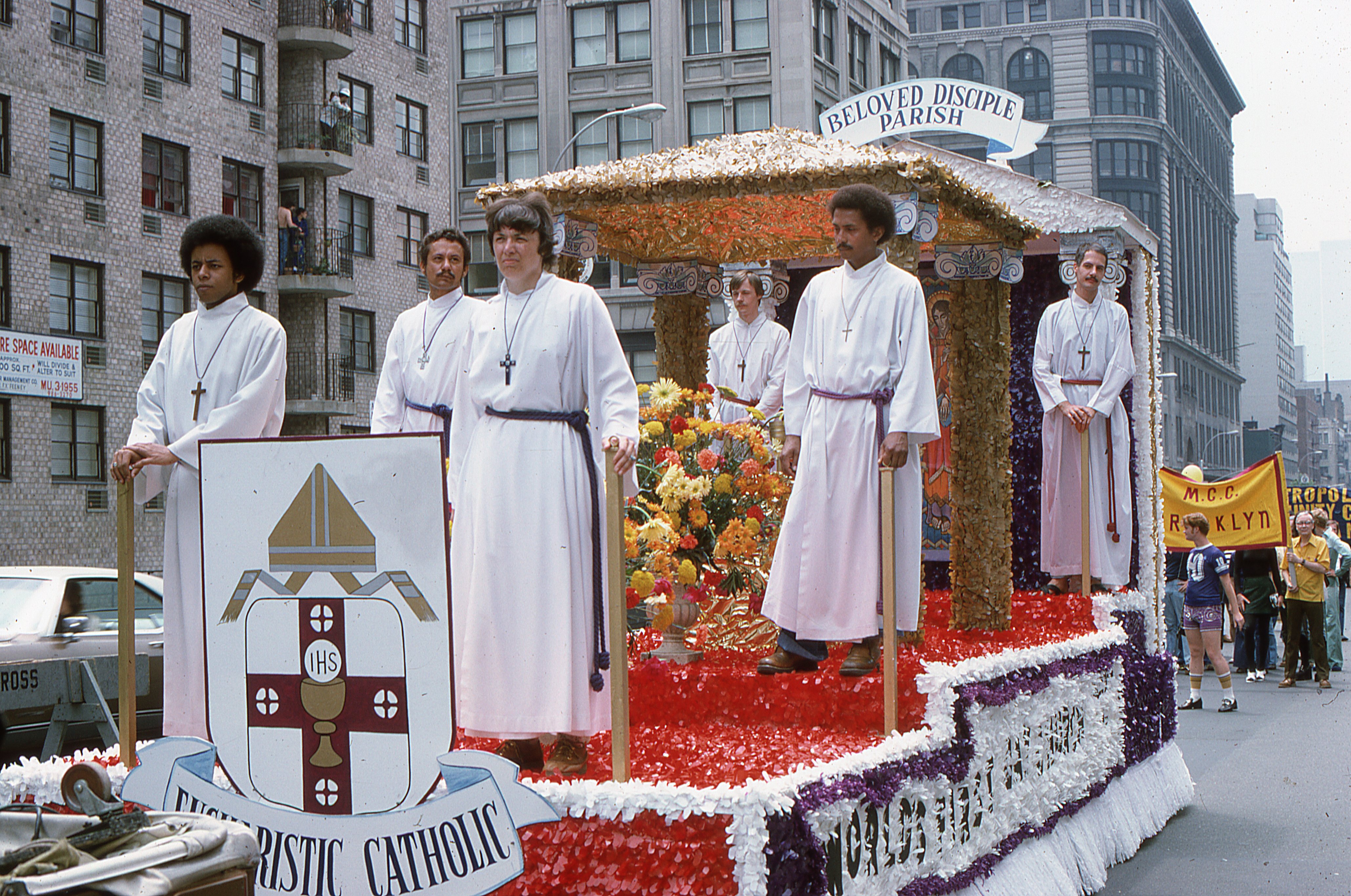 A closer view of the Church of the Beloved Disciple's float during the Sixth Annual Gay Pride Day March (Gay Liberation Day) in New York City on June 29, 1975. Decorated as a processional chapel and crowned with the sign "Beloved Disciple Parish," the float featured the congregation's heraldic arms and iconography, visually expressing its Catholic sacramental identity. The individuals on the float are presently unidentified, but their white albs and distinctive crosses reflected the liturgical character of the congregation's public witness.