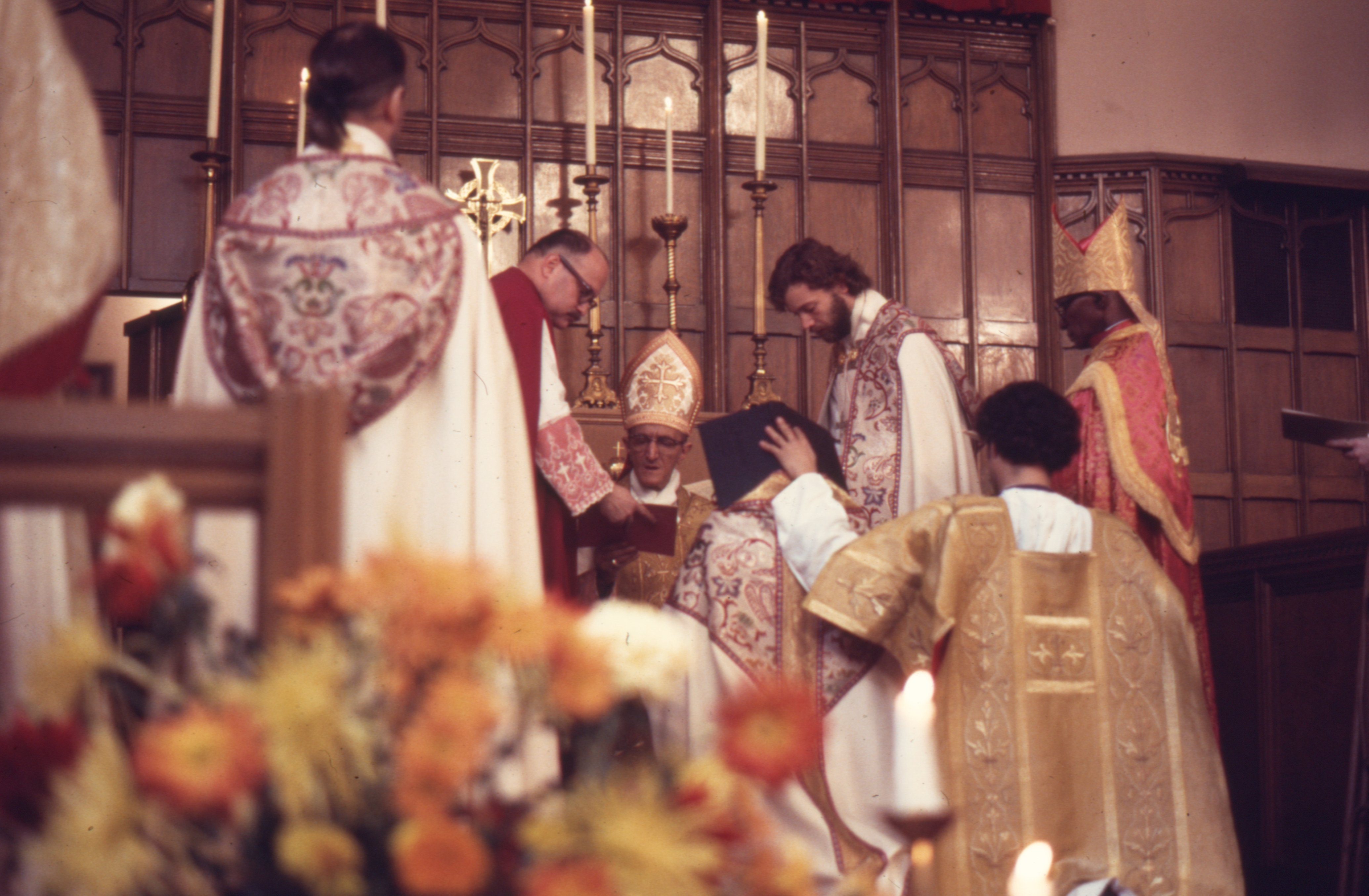 Robert Mary Clement kneels before the altar with the Book of the Gospels resting across his shoulders during the central rite of episcopal consecration at Metropolitan-Duane United Methodist Church in New York City on October 6, 1974. He was consecrated bishop by Archbishop Richard Arthur Marchenna, assisted by Bishop Martin Luther Williams, with Rev. Albert J. Berube of the North American Old Roman Catholic Church (Archdiocese of New York) also participating in the ceremony. At left, Fr. John Darcy Noble is seen from behind, while Fr. Leo Joseph stands to the right of Archbishop Marchenna. An unidentified acolyte supports the Gospel Book during the rite. Widely publicized and televised in New York City, the consecration was presented as the first public episcopal consecration of an openly gay bishop in the United States.