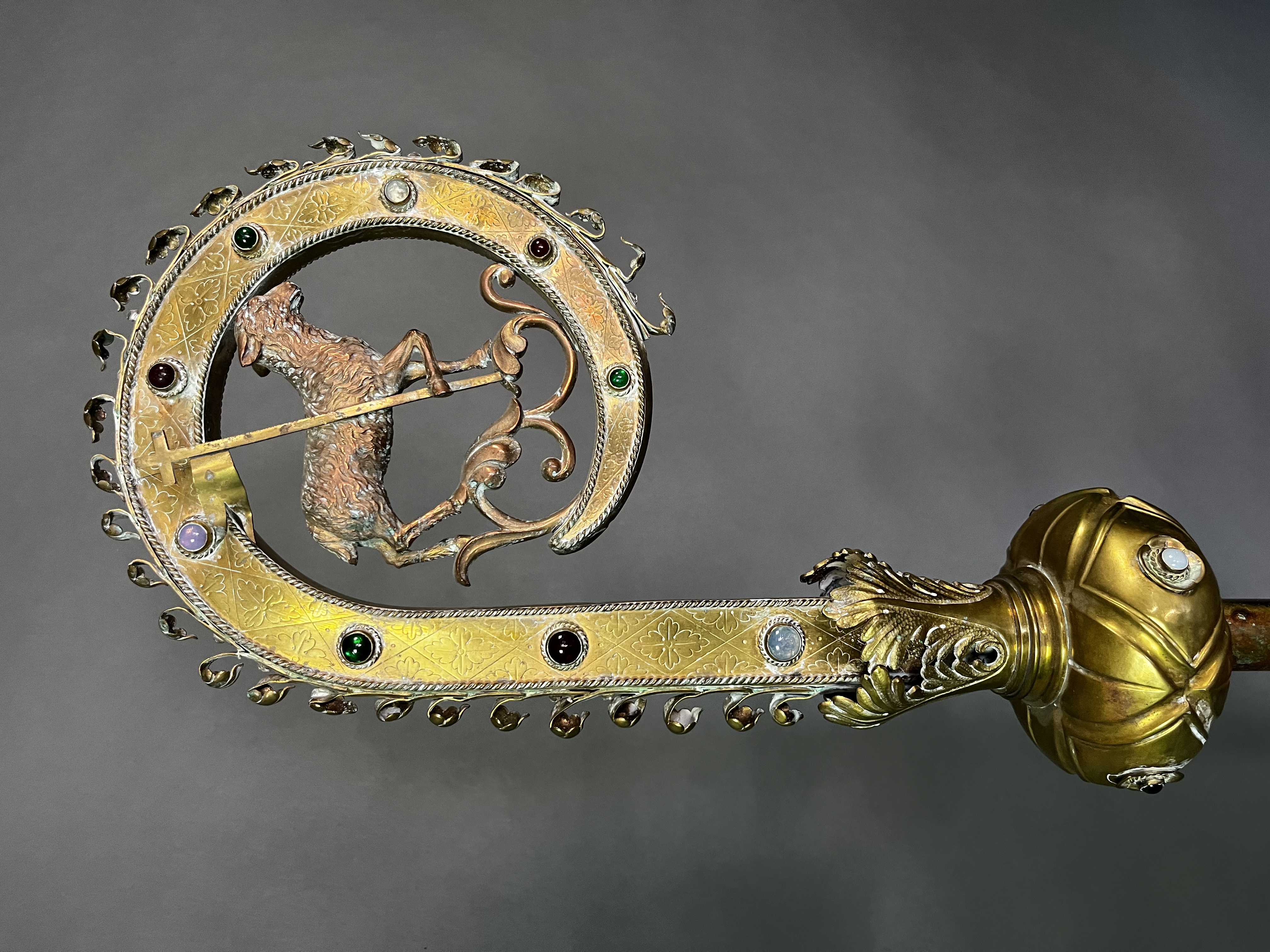 The crozier's elaborately worked volute centers on the Agnus Dei (Lamb of God) carrying the banner of the Resurrection, a traditional emblem of Christ's sacrificial death and triumph over death. The engraved brass is embellished with colored cabochons and scrolling foliate ornament, combining Gothic and Romanesque decorative motifs. As the principal symbol of the bishop's pastoral office, the crozier expressed Archbishop Clement's vocation as shepherd of the Church of the Beloved Disciple while proclaiming the resurrection hope that stood at the center of the community's sacramental life.