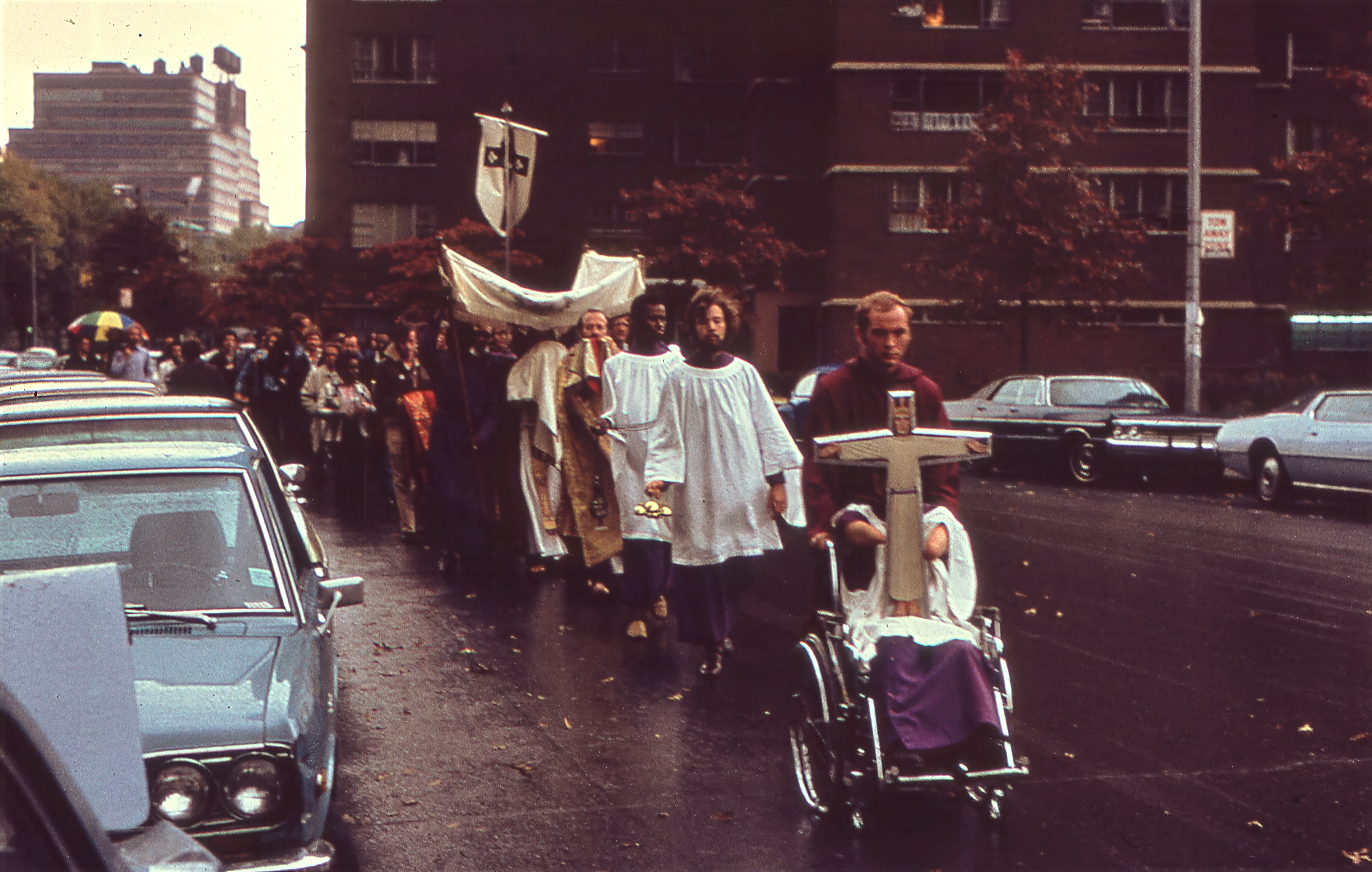 Led by a processional cross, acolytes, and a canopy sheltering the Blessed Sacrament, members of the Church of the Beloved Disciple process through the streets of Manhattan on the Feast of Corpus Christi. The annual procession carried the Eucharist beyond the church walls into the public square, affirming both the Real Presence of Christ and the place of LGBTQ Christians within the Catholic sacramental tradition. Rain and city traffic did not deter the congregation, whose public witness united ancient devotional practice with the visibility and resilience of New York's gay community.