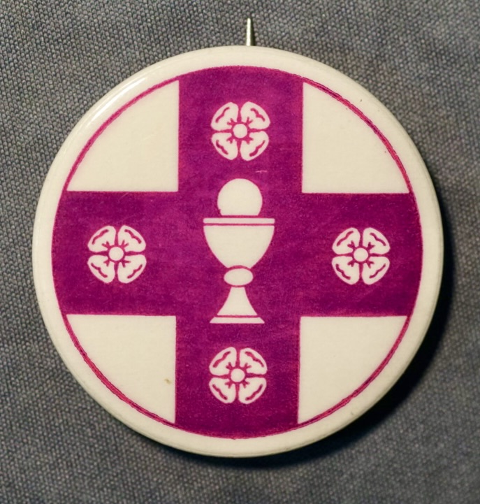 This small metal button reproduces the official emblem of the Church of the Beloved Disciple, presenting the congregation's distinctive visual identity without accompanying text. The design centers on a cross enclosing a Eucharistic chalice surmounted by the Sacred Host, symbolizing the centrality of the Eucharist in the church's theology and worship. Four stylized quatrefoils placed between the arms of the cross evoke both the four Evangelists and the universality of the Church. Distributed among members and supporters, the button was worn at liturgical celebrations, public processions, Pride marches, and ecumenical gatherings, serving as a recognizable symbol of the congregation's apostolic heritage and its public witness as one of the earliest openly LGBTQ-affirming sacramental churches in the United States.